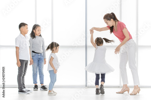 Asian woman in pink shirt dance with little Asian girl, Asian boy and gilrs look at them, they stand in front of big white window.