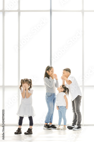 Asian kids point at Asian girl  the stand beside big white window.