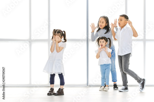 Asian kids point at Asian girl, the stand beside big white window.