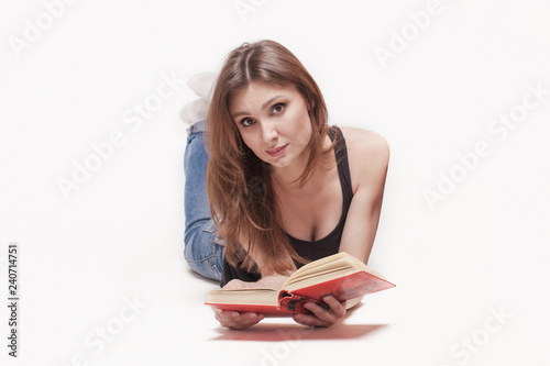 Young attractive caucasian woman lays on the floor legs up, book with red cover in the hands, slightly smile. Brown hair and eyes, wearing casual. White background, studio, isolated.