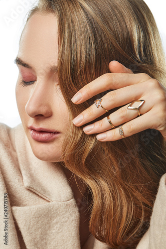 Closeup portrait of European lady with curly light brown hair, wearing a set of diversiform rings. The woman with rouge pink eyeshadow is touching her hair on white background with her eyes closed.  photo