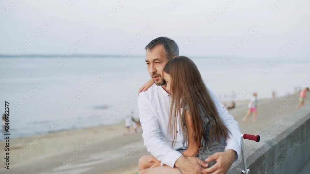 A girl sits on her father's lap. A man and his daughter are sitting on the embankment. Sunset. 4k A girl sits on her father's lap. A man and his daughter are sitting on the embankment. Sunset. 4k