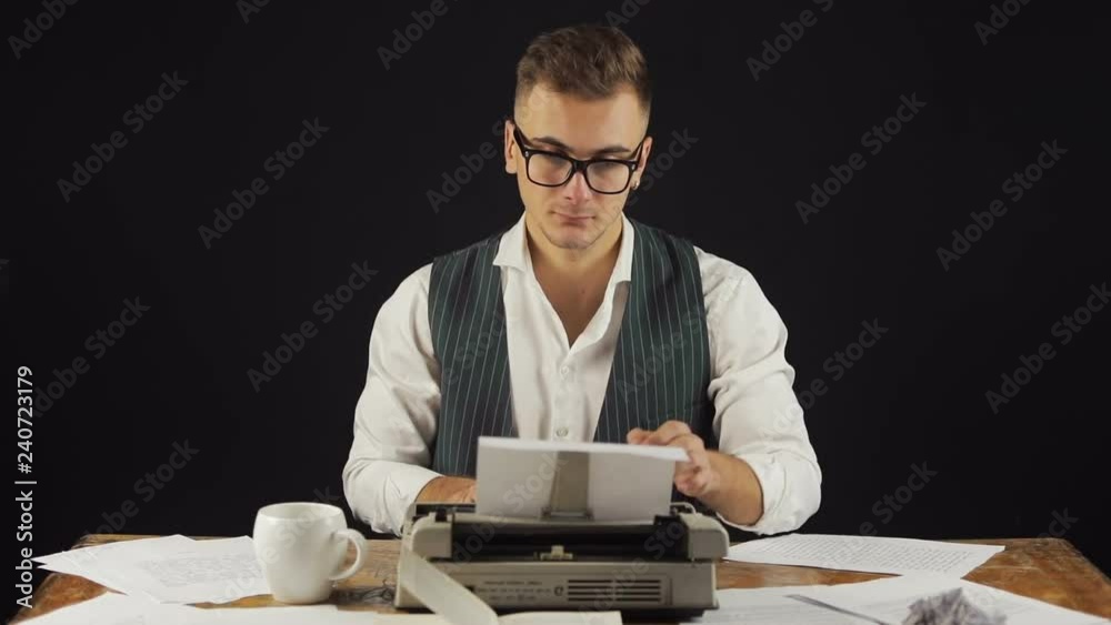 Serious writer finishes typing the text, clever fair-haired man in gray vest and glasses writing a novel on typewriter, indoor shoot in modern office with black walls Serious writer finishes typing the text, clever fair-haired man in gray vest and glasses writing a novel on typewriter, indoor shoot in modern office with black walls