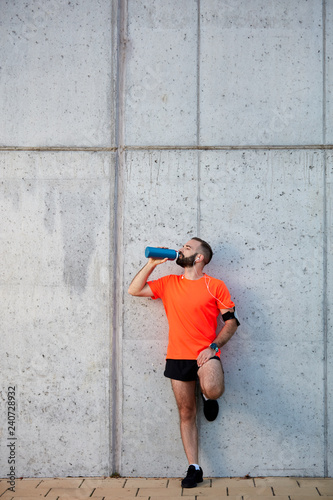 Man resting from running and drinking water while standing against the wall. Healthy lifestyle concept.