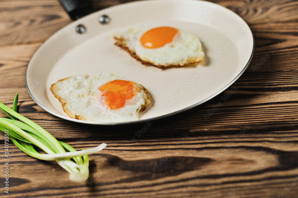 Two fried eggs in metal beige pan and green fresh onion on old wooden brown table. Copy space Two fried eggs in metal beige pan and green fresh onion on old wooden brown table. Copy space