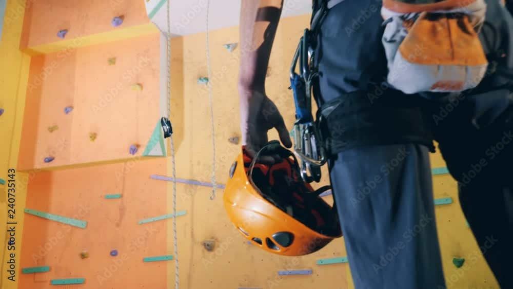 A man stands near a climbing wall with hardhat in hands, back view.