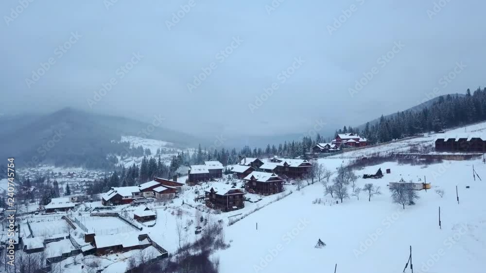 Aerial of inhabited locality in the mountains on winter. Mountain village buildings and houses on snowy hill slopes covered with snow. Countryside, fir tree and pine forest. Cottages of ski resort. Aerial of inhabited locality in the mountains on winter. Mountain village buildings and houses on snowy hill slopes covered with snow. Countryside, fir tree and pine forest. Cottages of ski resort.