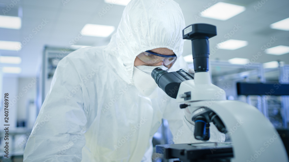 In Laboratory Scientist in Protective Clothes Doing Research Uses Microscope. Modern Manufactory Producing Semiconductors and Pharmaceutical Items. In Laboratory Scientist in Protective Clothes Doing Research Uses Microscope. Modern Manufactory Producing Semiconductors and Pharmaceutical Items.