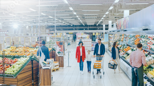 At the Supermarket: Happy Family of Three, Holding Hands, Walks Through Fresh Produce Section of the Store. Father, Mother and Daughter Having Fun Time Shopping. High Angle Panoramic Shot. photo