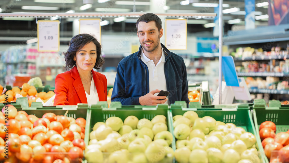 At the Supermarket: Happy Young Couple Chooses Organic Fruit in the Fresh Produce Section of the Store. Husband Uses Smartphone, Wife Picks Up Fruit. 