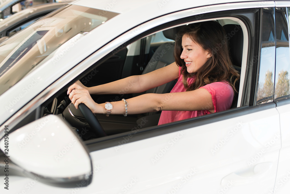 Happy woman buying a car. Beautiful smiling woman sitting and driving a car, hands on the wheel, in a car salon. Buying with cash or bank credit or leasing concept.