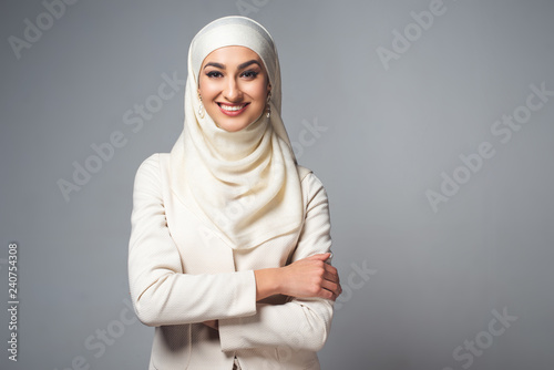 young muslim woman standing with crossed arms and smiling at camera isolated on grey photo