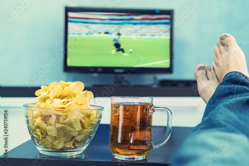 Closeup of male hand holding cup holding of beer with his feet crossed in front of a large television set sitting in his living room and watching football. fan watching the TV championship on channel. photo