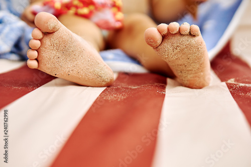 Close up of child's sandy feet on a colored towel sunbathe on the beach sunbed. Light heels of children's feet under the sun. The child is sunbathing, lying on the beach sunbed.