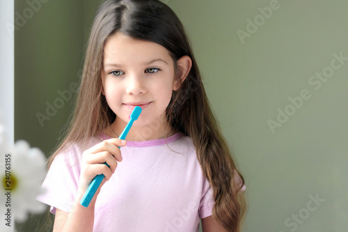 Long haired smiling beautiful caucasian white girl brushing her teeth with mint toothbrush on green background. Green eyed cute child in violet tshirt doing morning hygiene with blurred white flower 