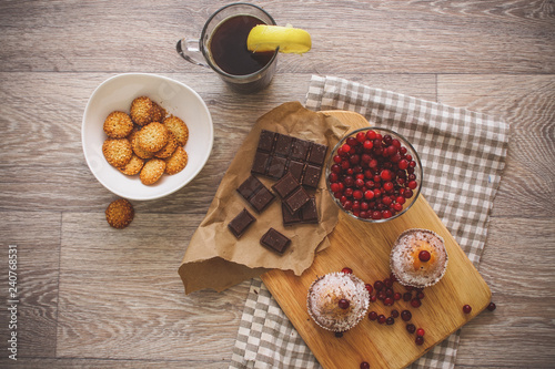 On a light wooden tabletop on a linen napkin napkin, there is a cutting board with two muffins, a broken chocolate bar and bright red berries in a small tree, next to a bowl with cookies.