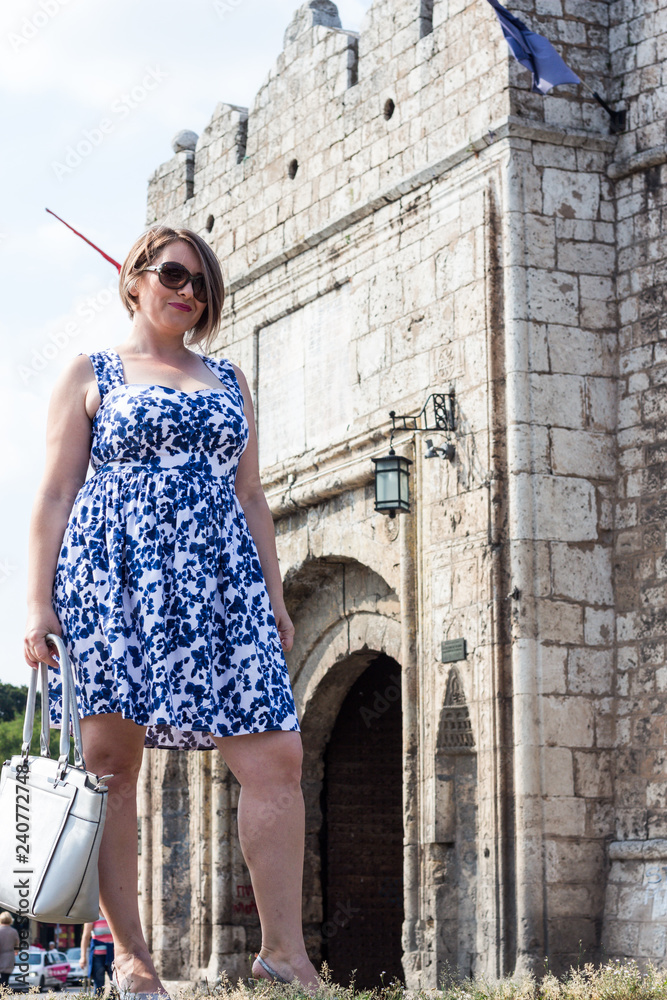 Beautiful Girl in a Blue Dress Holding Handbag and Standing in Front of the Fortress on a Beautiful Sunny Day in the City