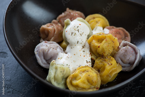 Boiled multicolored dumplings or pelmeni with sour cream in a black bowl, close-up, selective focus