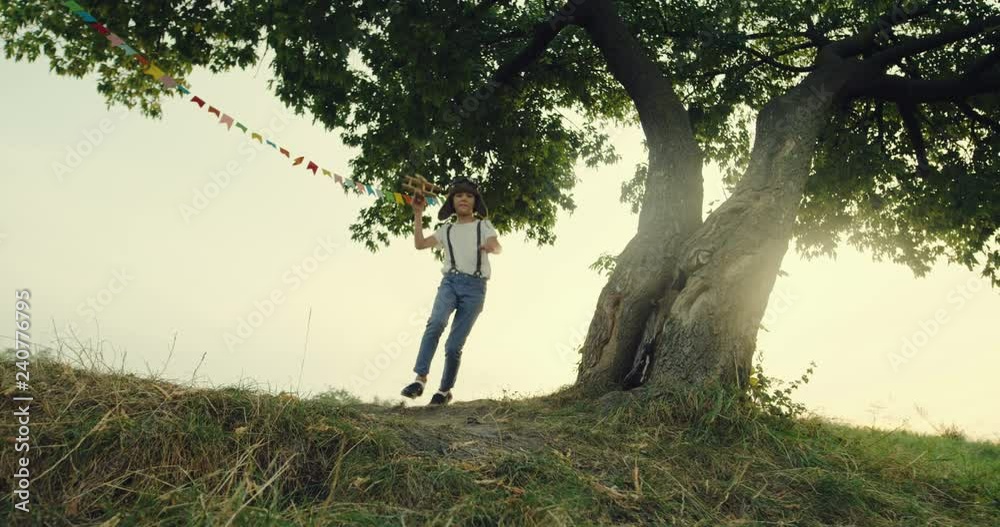 Caucasian teen boy dreamer of being a pilot in the special aviator hat and glasses running round the tree with a wooden plane in a hand. Caucasian teen boy dreamer of being a pilot in the special aviator hat and glasses running round the tree with a wooden plane in a hand.