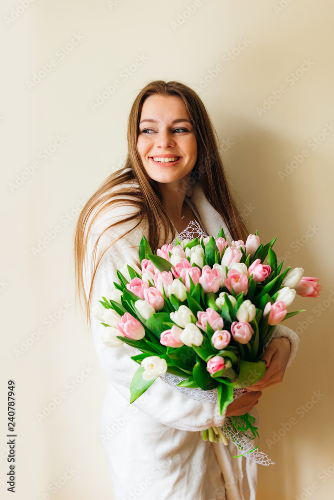 Joyful young woman in a dressing gown holds a bunch of fragrant pink and white flowers and smiles.