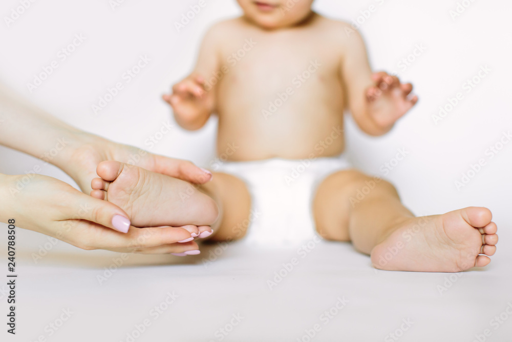 Mother massaging her child's foot, cute little baby girl in white diaper is sitting on the white background. Baby health and hygiene concept Mother massaging her child's foot, cute little baby girl in white diaper is sitting on the white background. Baby health and hygiene concept