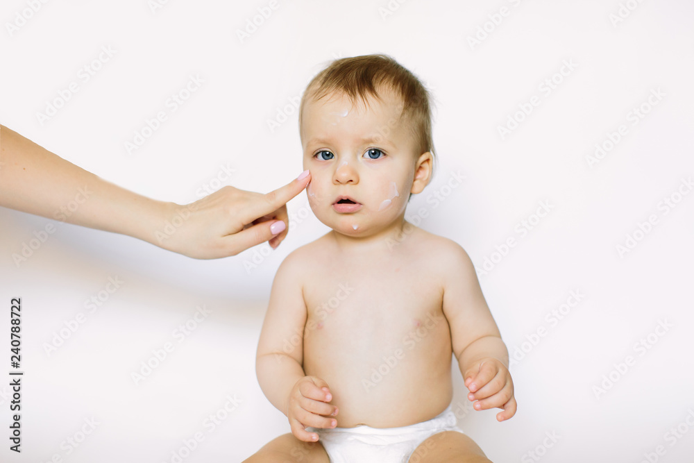Mother creaming cute infant girl face. Little baby girl with cream on her face sitting on the white background Mother creaming cute infant girl face. Little baby girl with cream on her face sitting on the white background