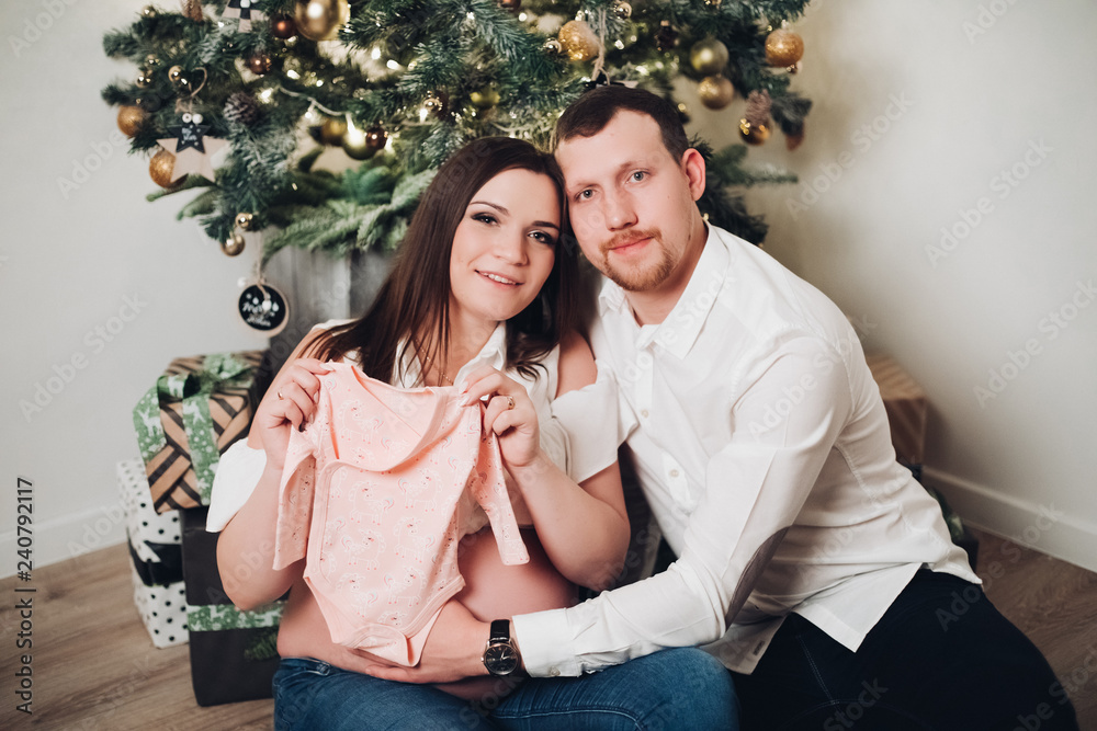 Happy couple sitting near christmas tree, looking at camera, smiling. Adorable woman holding baby cute bodysuit. Man wearing in white shirt holding hand on wife's belly.