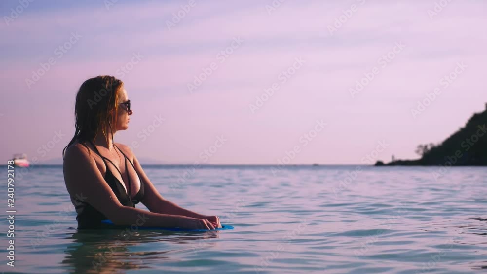 The girl the blonde in a black bathing suit and black glasses. A beautiful model with a sexy body swims holding a swimming board in crystal clear sea water.