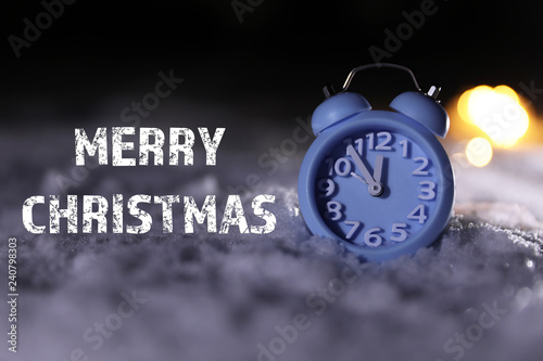 Blue alarm clock on snow and message MERRY CHRISTMAS against dark background. Winter holidays