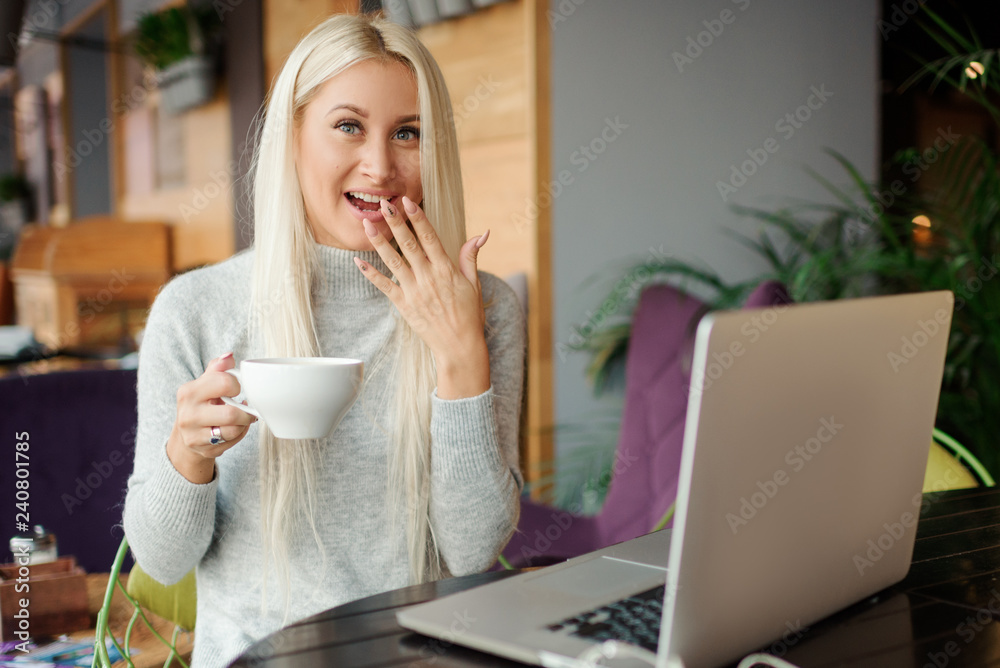 Shocked young woman in casual wear working on laptop while sitting in creative office or cafe and woking. Holding a cup of coffee and smiling Shocked young woman in casual wear working on laptop while sitting in creative office or cafe and woking. Holding a cup of coffee and smiling