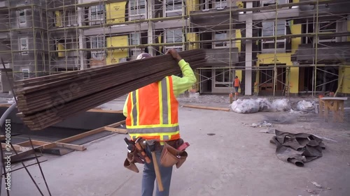 A construction worker walking and working on a construction site wearing a tool belt and high visibility clothing carrying lumber. 