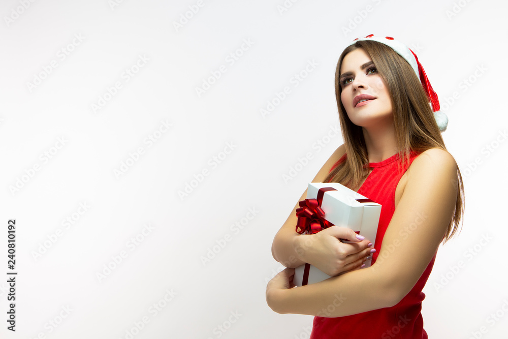 Christmas Time Concepts. Happy Calm Caucasian Female In Red Dress and Santa Hat. Posing With White Gift Box Wrapped Up with Red Ribbon. Isolated Over White. Christmas Time Concepts. Happy Calm Caucasian Female In Red Dress and Santa Hat. Posing With White Gift Box Wrapped Up with Red Ribbon. Isolated Over White.