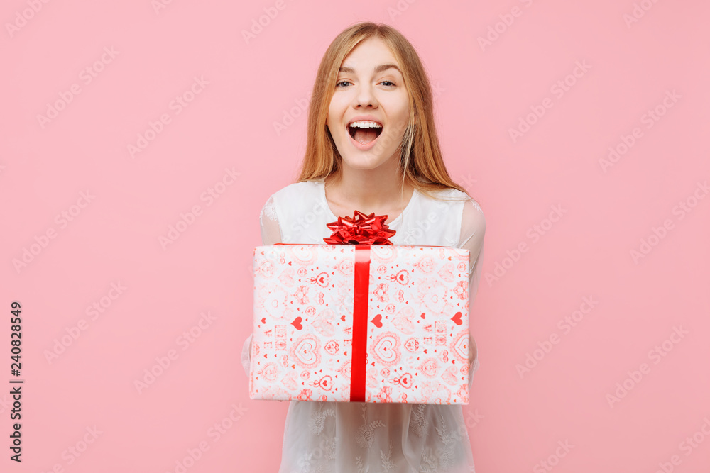 Shocked young woman, in a white dress, with a gift box in her hand, delighted with the gift, on a pink background Shocked young woman, in a white dress, with a gift box in her hand, delighted with the gift, on a pink background
