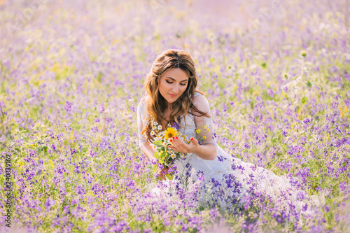 lovely sweet woman with closed eyes dressed in an elegant white dress with transparent sleeves sits in purple flowers with a natural field bouquet in her hands. atmospheric photo, gentle warm colors
