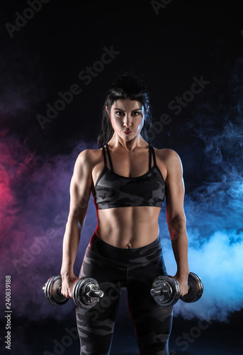 Sporty muscular woman with dumbbells in smoke on dark background