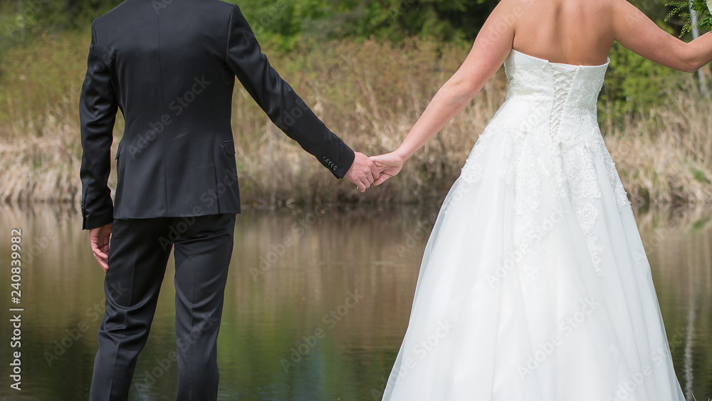 Hochzeitspaar in schönen Kleidern steht vor einem Teich und hält sich an den Händen