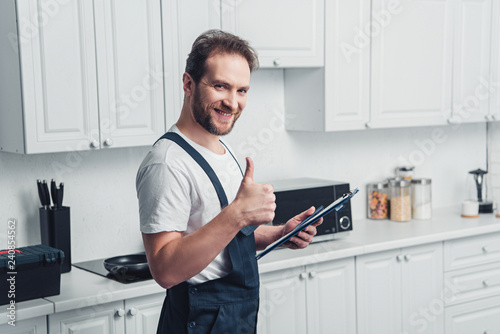 smiling handsome adult bearded repairman holding clipboard and doing thumb up gesture in kitchen