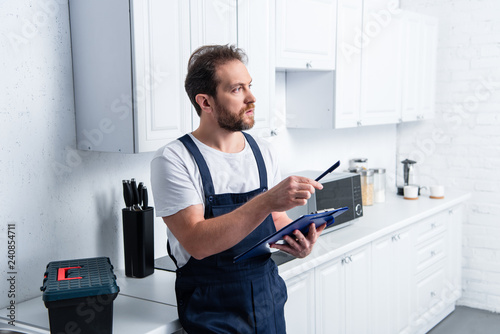 male bearded craftsman pointing by pen and holding clipboard in kitchen