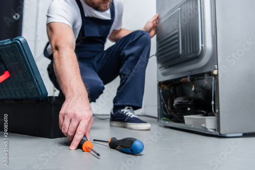 фотографія cropped shot of repairman in working overall taking screwdriver from floor near