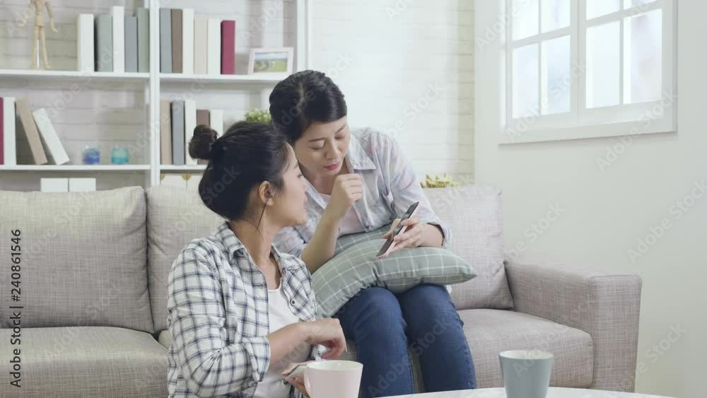 two happy friends sharing mobile screen discussing touching choosing joyfully. asian girls sharing online content with multiple devices sitting on a sofa at home in afternoon tea time indoors. two happy friends sharing mobile screen discussing touching choosing joyfully. asian girls sharing online content with multiple devices sitting on a sofa at home in afternoon tea time indoors.