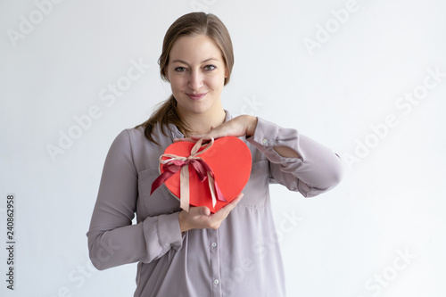 Content pretty young woman showing heart shaped gift box. Girl wearing casual shirt and looking at camera. Saint Valentines Day gift concept. Isolated front view on white background.