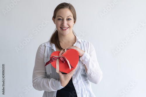 Happy pretty young woman holding heart shaped gift box. Girl looking at camera. Saint Valentines Day gift concept. Isolated front view on white background.