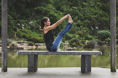 Young woman in urban clothing performing ubhaya padangusthasana on park bench by pond. Female yogi in balancing stick pose grabbing toes with hands photo