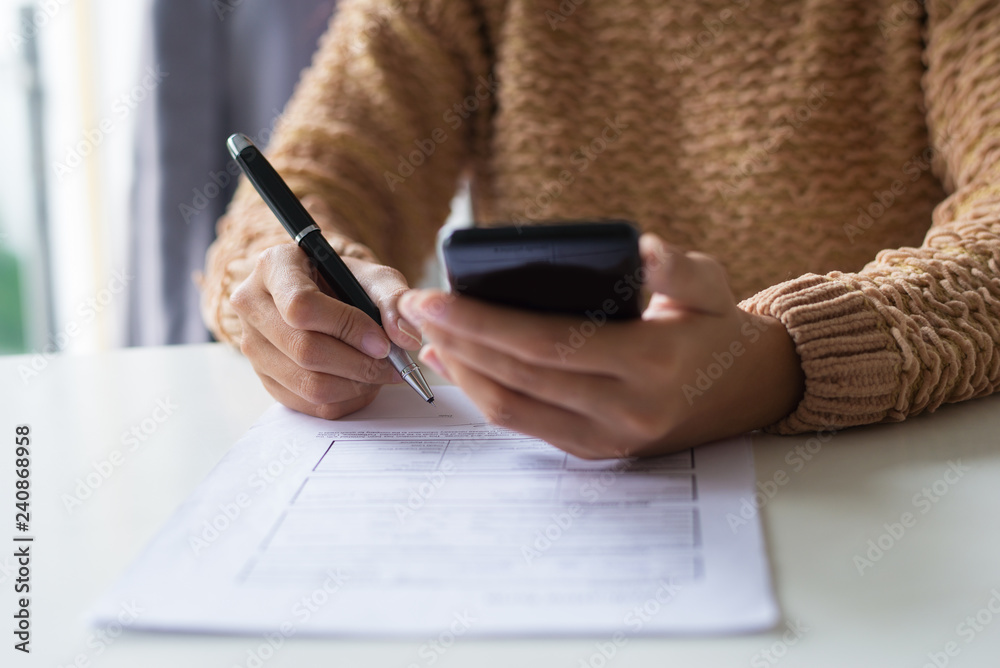 Close-up of lady in sweater checking message on smartphone. Unrecognizable woman filling tax form. Paperwork concept