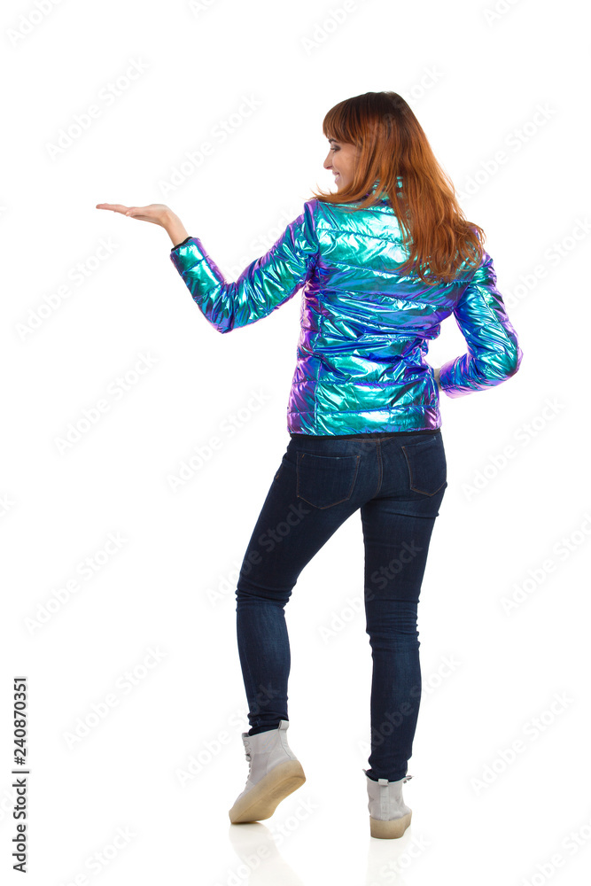 Young Woman In Vibrant Down Jacket Is Holding Hand Raised And Showing Something. Rear View. Young Woman In Vibrant Down Jacket Is Holding Hand Raised And Showing Something. Rear View.