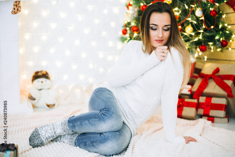 girl in white sweater and jeans sitting on a Christmas tree background, tender and cute