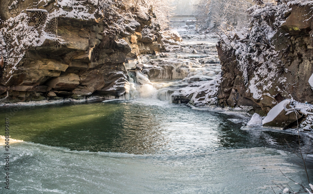 Naklejka premium icy winter waterfall steaming between stones, orange sunlight, frozen pond