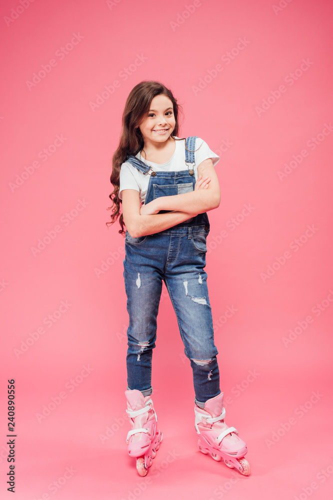 Naklejka premium happy child in overalls with arms crossed wearing rollerblades on pink background