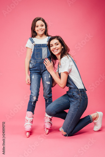 happy mother hugging daughter in roller blades and looking at camera on pink background