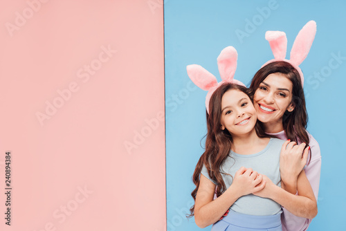 Canvas Print beautiful woman in bunny ears hugging child and smiling at camera on blue and pi